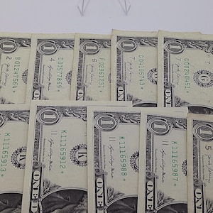 May include: Several United States one-dollar bills are arranged on a white surface. The bills display the portrait of George Washington and various serial numbers. The bills are green and black.