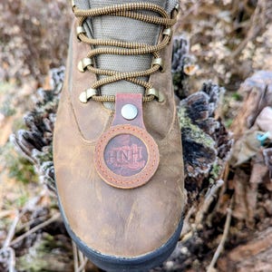May include: Close-up of a brown leather hiking boot with olive green accents and brown laces. A circular leather tag with the letters "HNH" is attached to the laces. The boot is set on a natural, outdoor background.