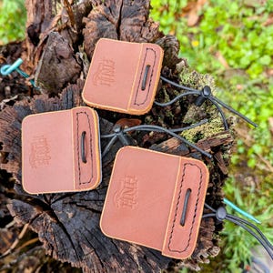 May include: Three brown leather pouches with black drawstrings are displayed on a weathered tree stump. Each pouch has a rectangular shape with rounded corners and a stitched border. The logo "HNH" is embossed on each pouch.