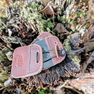 May include: Leather keychains with a two-tone brown design and a black cord. One keychain has the initials "ANH" embossed on it. The keychains are displayed on a weathered tree stump.