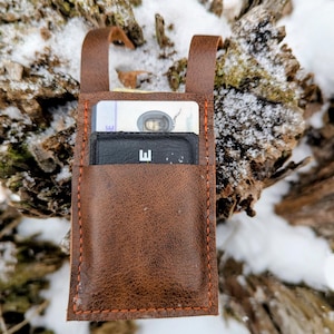 May include: A brown leather card holder with orange stitching. The card holder has two loops at the top and contains several cards. The background is a blurred view of a snow-covered tree trunk.