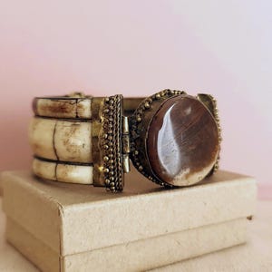 May include: A vintage bracelet with a large, polished brown stone set in a brass-toned metal frame. The bracelet has three ivory-coloured segments and decorative metal accents. The bracelet is displayed on a small, beige box.