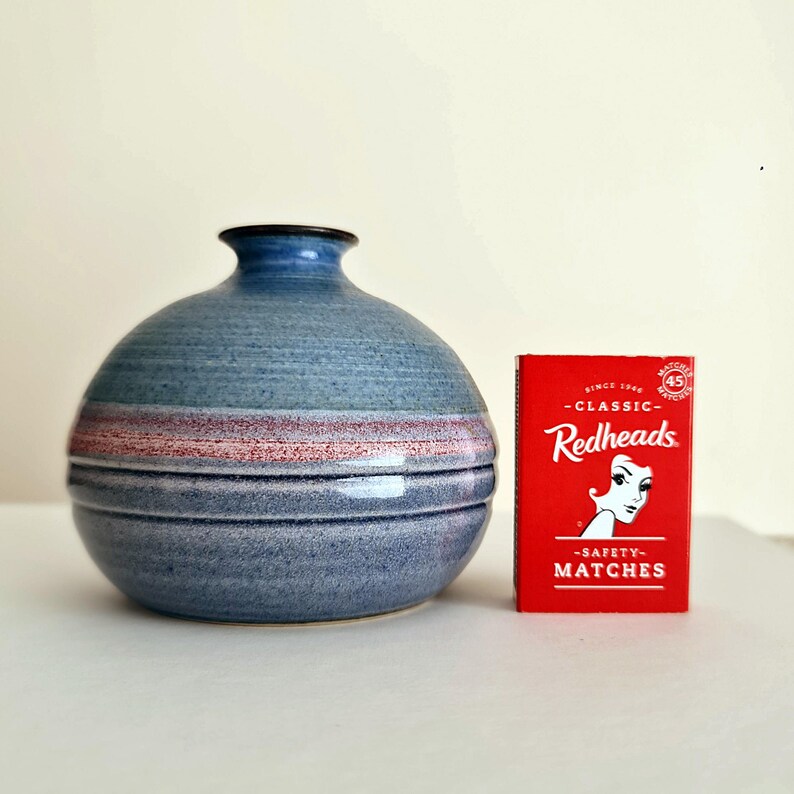 Pu&ograve; includere: Un vaso rotondo in ceramica blu con un bordo blu scuro e una fascia orizzontale di strisce rosa e rosse. Una scatola di fiammiferi rossa con il testo "Redheads" e "Safety Matches" &egrave; accanto al vaso.