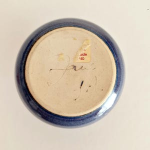 Pu&ograve; includere: Una ciotola di ceramica blu con interno beige chiaro. Il fondo della ciotola presenta firme manoscritte e un adesivo con la scritta "aide '40". La ciotola ha una forma rotonda e una finitura lucida.