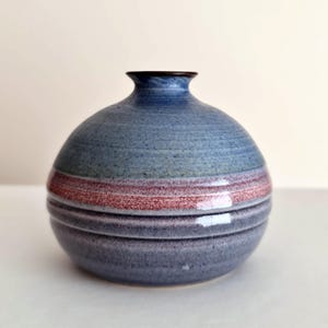 May include: A round ceramic vase with a blue and red striped design. The vase has a dark brown rim and a textured surface. The vase is approximately 15 cm tall and 15 cm wide.