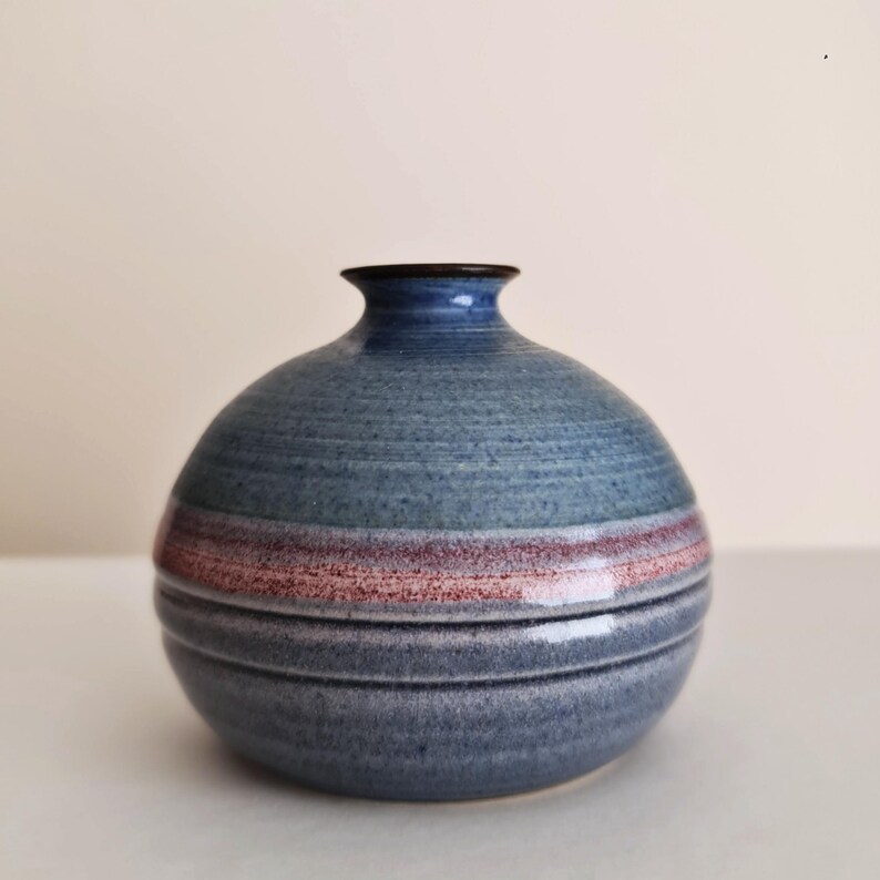 Pu&ograve; includere: Un vaso rotondo in ceramica blu con collo stretto e bordo scuro. Il vaso presenta fasce orizzontali di colore, tra cui una striscia rosso-marrone al centro. Il vaso &egrave; un oggetto decorativo, adatto per esporre fiori o come opera d'arte a s&eacute; stante.