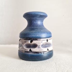 May include: A blue ceramic vase with a decorative band of white and grey geometric cutouts. The vase has a flared top and a rounded base. The colour is a deep, matte blue with a textured finish, giving it a handcrafted appearance.