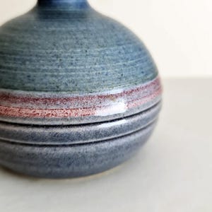 Pu&ograve; includere: Un piccolo vaso rotondo in ceramica con superficie strutturata. Il vaso &egrave; principalmente blu-grigio con una fascia orizzontale rossa e rosa. La base presenta diverse linee orizzontali grigio scuro. Il vaso ha un collo stretto e un corpo arrotondato.
