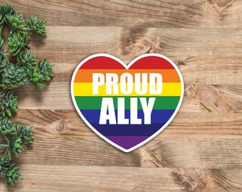 May include: A heart-shaped sticker featuring horizontal stripes in red, orange, yellow, green, blue, and purple. The text PROUD ALLY is printed in bold white capital letters across the center of the rainbow-colored heart design.