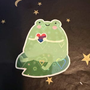May include: A die-cut sticker featuring a light green cartoon frog sitting on a dark green lily pad. The frog has pink cheeks and holds a small heart with horizontal stripes in pink, purple, and blue. The sticker has a holographic finish.
