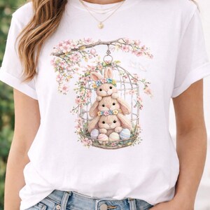 May include: White t-shirt featuring a whimsical Easter design. The graphic depicts three adorable bunny rabbits inside a birdcage, surrounded by pastel-colored Easter eggs and blooming flowers. A charming spring-themed apparel item.