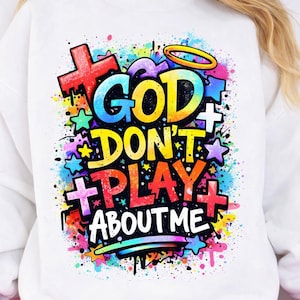 May include: White sweatshirt with a colorful graphic that reads "GOD DON'T PLAY ABOUT ME." The design includes crosses, stars, and a halo, with a vibrant, multi-colored splatter effect. The text is in bold, eye-catching fonts.