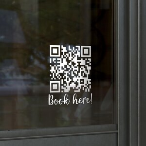 May include: A glass door featuring a QR code sticker and the text "Book here!". The QR code is black and white, and the text is in a cursive font. The door has a silver frame and a gold handle.
