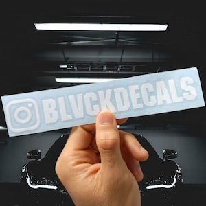 May include: A rectangular white sticker with the text "BLVCKDECALS" in a bold, sans-serif font. The Instagram logo is on the left. The sticker is held in front of a black car.