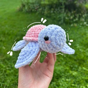 May include: A handmade crochet turtle in soft pastel colors. The turtle has a pink shell, a light blue head and flippers, and black eyes. The plush toy is designed with a cute, friendly expression, perfect for a child's toy or nursery decor.