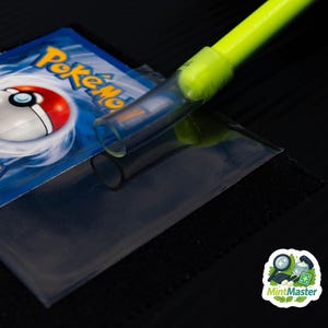 May include: A close-up of a Pokémon trading card being inserted into a clear plastic sleeve. A neon green and clear tool is used to assist with the insertion. The Pokémon card has a red and white Pokéball design and the word "Pokémon" in yellow.