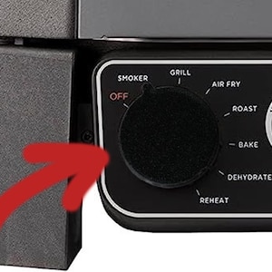 May include: A close-up of a black control panel with a dial and cooking function labels. The dial has settings for smoker, grill, air fry, roast, bake, dehydrate, reheat, and off. The panel also features a Woodfire Technology logo.