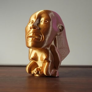 Puede incluir: Escultura dorada de una figura humana con la boca abierta y rasgos faciales detallados. La escultura tiene un diseño estilizado y está sobre un fondo neutro. La figura parece estar arrodillada.