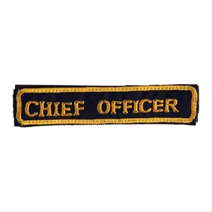 May include: A rectangular patch with the words "CHIEF OFFICER" embroidered in gold thread on a navy blue background. The patch has a gold border and is likely for a uniform.