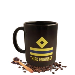 Peut inclure: Mug à café noir avec un insigne jaune et bleu et l'inscription "THIRD ENGINEER". Une cuillère en bois remplie de grains de café repose sur un lit de grains de café à côté de la tasse.