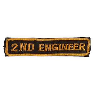 May include: A black rectangular patch with gold embroidered text reading "2ND ENGINEER". The patch features a gold border. The text is in a bold, sans-serif font. It is designed to be sewn onto clothing or equipment.