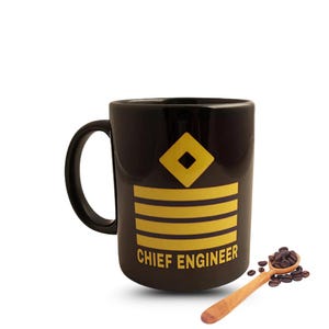 Peut inclure: Mug en céramique noire avec un motif d'ingénieur en chef. La tasse présente un losange et des rayures jaunes, avec les mots "CHIEF ENGINEER" en jaune. Une cuillère en bois avec des grains de café est à côté de la tasse.