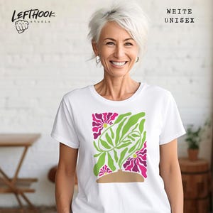 May include: White unisex t-shirt featuring a floral design with pink and green elements. The design includes stylized flowers and leaves, set against a brown vase. The shirt has the text "WHITE UNISEX".