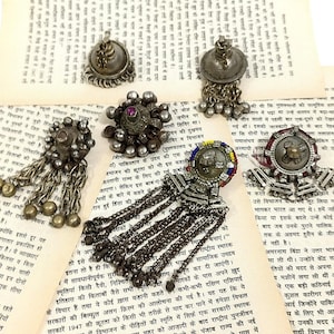 Può includere: Una collezione di sei ciondoli in argento e metallo con disegni intricati e catene pendenti. I ciondoli presentano una varietà di forme, tra cui rotonde, quadrate e a forma di stella. Alcuni dei ciondoli hanno piccole perline o pietre attaccate.