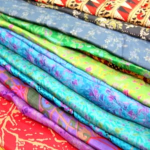 May include: A stack of colorful patterned fabric scarves in various shades of blue, green, purple, and red. The scarves are folded and layered on top of each other, creating a visually appealing display.