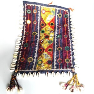 Puede incluir: Textil rectangular hecho a mano con un diseño de patchwork. El panel central presenta patrones geométricos en amarillo, rojo y morado, adornado con pequeños espejos. Los bordes están adornados con conchas blancas y borlas.