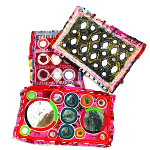 May include: Three decorative fabric pieces with mirror details. The pieces are in red, pink, and black, featuring intricate embroidery and mirrors of different sizes. The edges are trimmed with colourful threads. The pieces are likely handmade.
