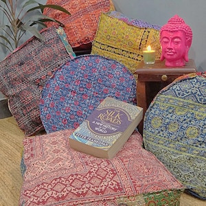 May include: A collection of colorful, patterned floor cushions with a book titled "The Silk Roads: A New History of the World" by Peter Frankopan on top of one of the cushions. The cushions are arranged on a woven rug with a pink Buddha statue and a candle on a wooden table in the background.