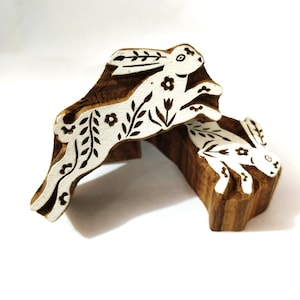 May include: Two wooden block stamps with a rabbit design. The rabbits are white with floral and leaf details, set against a brown wooden background. The stamps are for printing or crafting.