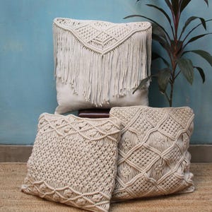 May include: Three decorative throw pillows with a macrame design. The pillows are made of natural cotton rope and have a fringe detail. The pillows are arranged on a light brown surface.