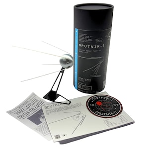 May include: A Sputnik-1 model kit with a black cylindrical container. The kit includes a model of the satellite, a stand, and printed materials. The satellite is silver with wire-like antennas. The container has the text "SPUTNIK-1".