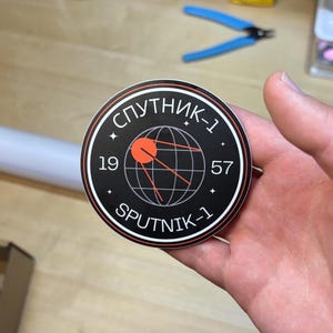 May include: A round black sticker with white and red accents. The sticker features the text "СПУТНИК-1" and "SPUTNIK-1" in white, along with the numbers "19" and "57". A red graphic of a satellite and a globe is in the center.