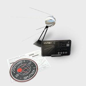 May include: A silver Sputnik 1 model with four antennae, displayed on a black stand. A black information card and a circular sticker with Cyrillic text and a red dot are also present. The model is a replica of the first artificial satellite.
