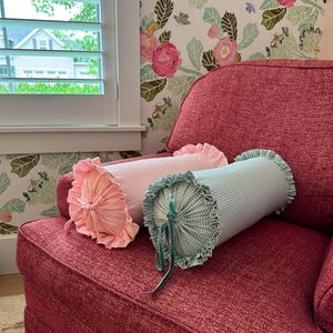 May include: Two cylindrical bolster cushions on a red armchair. One is pale pink with frilled edges, the other is light blue with a gingham pattern and green velvet ties. Floral wallpaper and a window are in the background.