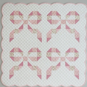 Può includere: Una coperta trapuntata per bambini con un motivo ripetuto di fiocchi rosa su sfondo bianco. I fiocchi sono realizzati in tessuto rosa e a fantasia floreale. La coperta ha un bordo smerlato con un bordo rosa.