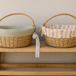 Peut inclure: Deux paniers en osier avec des doublures en tissu. Le panier de gauche a une doublure à carreaux bleu clair et blanc. Le panier de droite a une doublure à motifs floraux avec des couleurs blanches, jaunes et roses. Les deux paniers sont posés sur une étagère en bois.