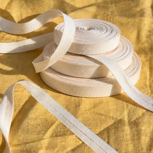 May include: Three stacked rolls of off-white cotton bias tape, each featuring a single line of stitching running down the center. The tape is unspooled, with a long length extending outward from the stack, showcasing the fabric texture and color.
