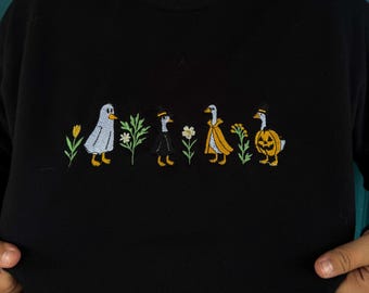 May include: Black sweatshirt with embroidered Halloween geese. The geese are dressed in costumes: a ghost, a witch, a princess, and a pumpkin. The design includes flowers and is centered on the chest.