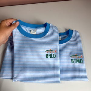May include: Two short-sleeved t-shirts featuring a light blue and white horizontal striped pattern with a solid blue crew neckline. Each shirt displays an embroidered graphic of a fish above the green embroidered letters BNLD or BTMD.