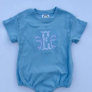 May include: A light blue baby romper with short sleeves and a round neckline. The romper features an embroidered monogram in white and light blue. The fabric is soft and gathered at the bottom with snap closures.