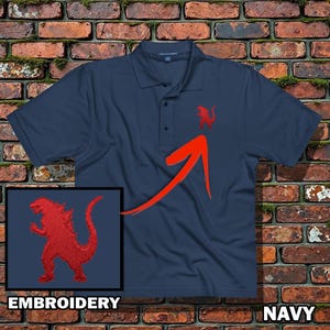 May include: Navy blue polo shirt with a red embroidered Godzilla design on the chest. The shirt features a classic collar and button closure. The word "EMBROIDERY" is displayed in a box at the bottom left, and the word "NAVY" is displayed at the bottom right.