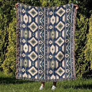 May include: A woven throw blanket with a geometric pattern in navy blue, cream, and olive green. The design features diamond and zigzag shapes. The blanket has fringed edges and is held up against a backdrop of green foliage.