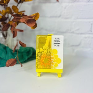 May include: Yellow glass block with a white background and a yellow easel stand. The glass block has a yellow and orange mandala design and the text "Be the reason someone smiles!"