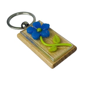 May include: A wooden keychain with a blue flower and green leaves embedded in clear resin. The keychain has a silver metal ring.