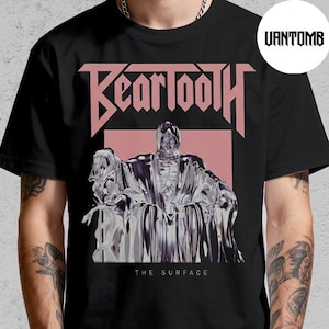 May include: Black t-shirt featuring the band name "Beartooth" in a pink, stylized font above a graphic of a figure in a robe. The text "THE SURFACE" is below the graphic. The shirt is worn by a person.