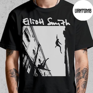 May include: Black t-shirt featuring a black and white graphic of figures falling from a building, with the text "Eliott Smith" in white. The shirt is a crew neck style.